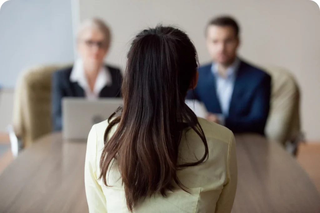 3 people sitting at a conference table, one with their back to the camera, and the other two facing forward. The two appear to be interviewing the one.